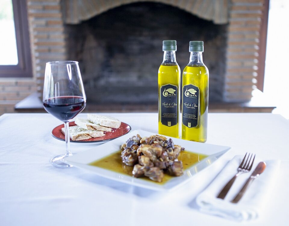 Mesa junto a una chimenea. Sobre la mesa una copa de vino, cubiertos, plato con pan, plato con carne de cordero y destacando dos botellas de medio litro de AOVE Flor de Oro.